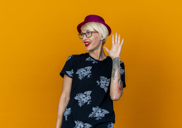 衣服令人印象深刻的年轻金发党妇女戴着党的帽子和眼镜在一旁做嗨手势孤立的橙色墙上姿势站着感觉