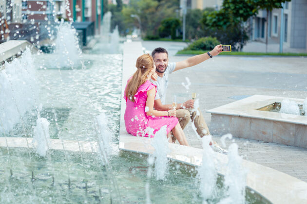 男朋友美丽的地理位置可爱的年轻夫妇坐在喷泉边自拍约会感情设备