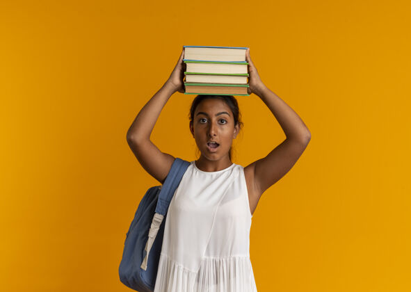 背惊讶的年轻女学生戴着背袋拿着书在橙色的头上女学生抱年轻