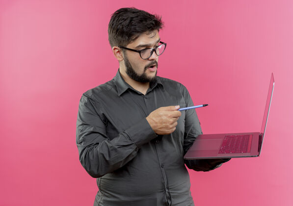 穿印象深刻的年轻商人戴着眼镜拿着笔指着隔离在粉红色墙上的笔记本电脑年轻印象笔
