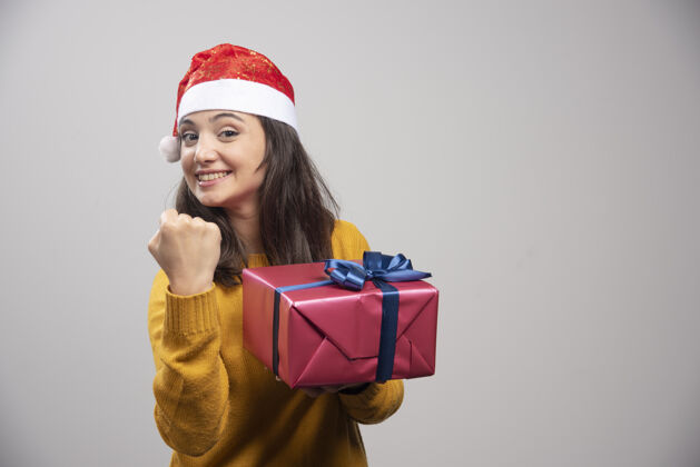 庆祝戴着圣诞帽的黑发女人伸出拳头 手里拿着礼盒表情圣诞节人