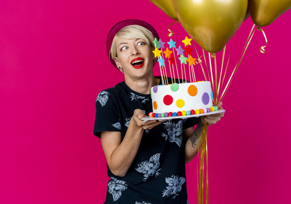 市民令人印象深刻的年轻金发派对女郎戴着派对帽拿着气球和生日蛋糕与明星看着蛋糕隔离在深红色的墙壁女人生日人