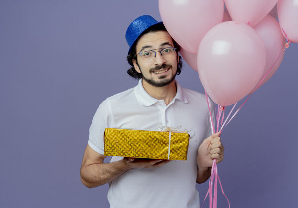 男人笑容可掬的帅哥戴着眼镜 戴着蓝色的帽子 手里拿着气球和礼盒 孤立在紫色上气球帅气微笑
