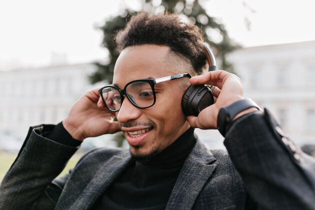 快乐在街上听音乐的有趣而惊讶的家伙的特写肖像戴眼镜的非洲男人戴着耳机摆姿势年轻成人音乐
