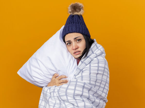 帽子虚弱的年轻白种人生病的女孩戴着冬天的帽子和围巾 裹在格子布抱枕里 看着孤立在橙色背景上的相机 还有复制空间枕头围巾疾病