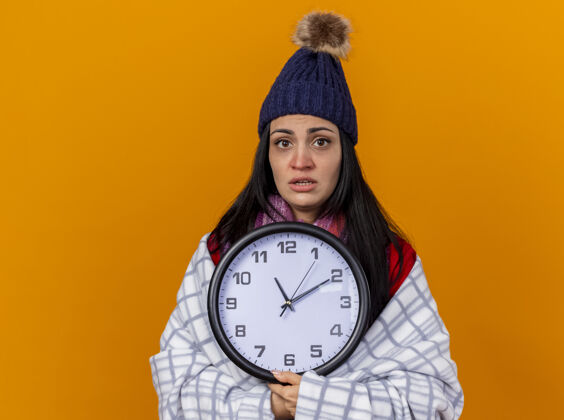 橙色印象深刻的年轻白种人生病的女孩戴着冬天的帽子和围巾包裹在格子布拿着时钟看着相机孤立在橙色背景与复制空间围巾时钟年轻