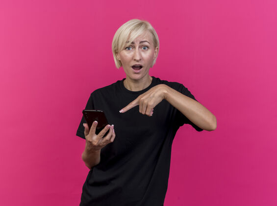 手机印象深刻的中年金发女子拿着手机指着前面孤立的粉色墙壁脸姿势衣服