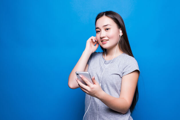 人亚洲年轻女子通过耳机听有声读物在蓝墙上特写年轻情感