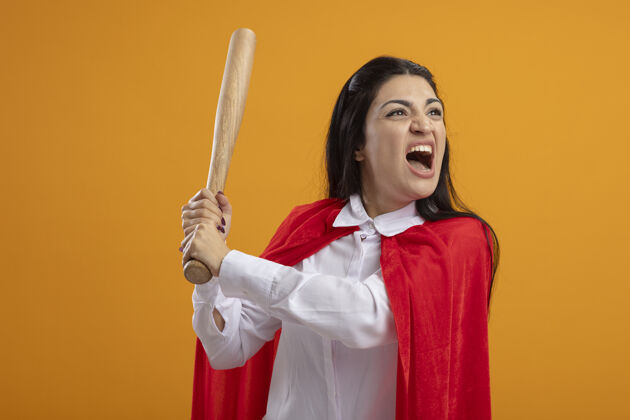 愤怒愤怒的年轻女超人拿着棒球棒看着橙色墙上孤立的一面女人棒球立场