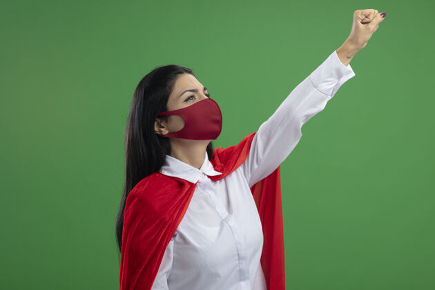 公民自信的年轻女超人戴着面具站在侧视图举起她的拳头像超人看隔离在墙上面具信心人