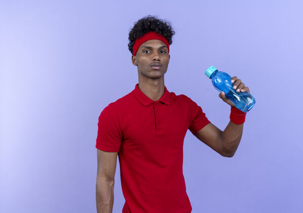 男人自信的年轻美国黑人运动男子戴着头带和腕带拿着水瓶隔离在蓝色的墙上年轻运动墙