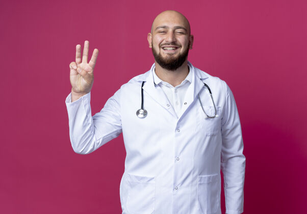 医生高兴的年轻秃头男医生穿着医用长袍和听诊器显示三个孤立的粉红色墙上长袍请表演