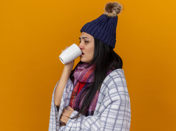 女人愁眉不展的年轻病女人戴着冬天的帽子和围巾裹着格子呢站在侧视图喝着茶看起来直孤立在橙色的墙上表情衣服轮廓