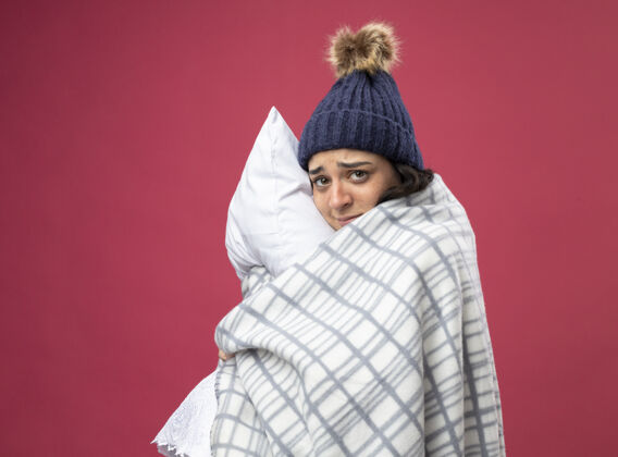 疾病体弱多病的年轻女子穿着长袍 戴着冬天的帽子 围着方格呢围巾站在侧视图里 抱着枕头 看着隔离在粉色墙上的前面人表情女人