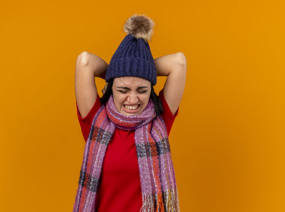 帽子带着冬日帽子和围巾的疼痛的生病的年轻女人 双手放在脖子后面 闭着眼睛 孤立地站在橙色的墙上女人穿着感觉