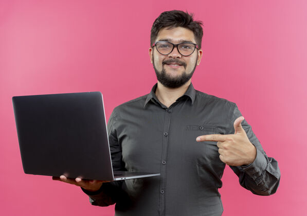 请高兴的年轻商人戴着眼镜 指着隔离在粉红色墙上的笔记本电脑穿商务人士年轻人