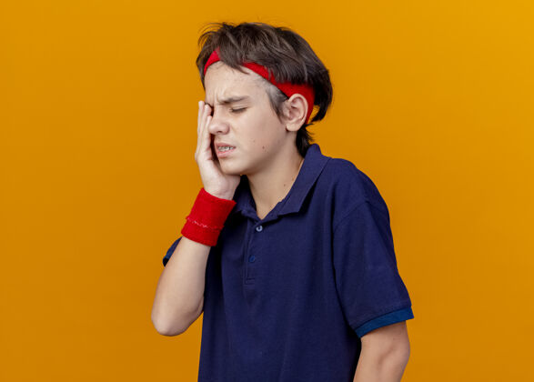 背带疼痛的年轻英俊的运动男孩戴着头带和护腕 戴着牙套 手放在橙色墙上的眼睛上立场衣服眼睛