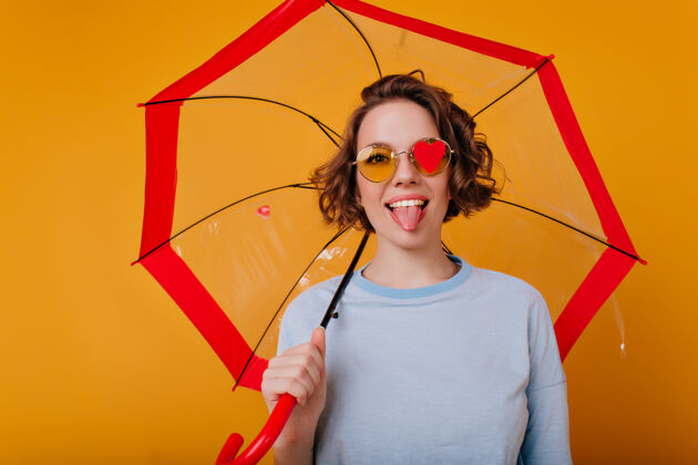 黑发优雅的年轻女模特伸着舌头 站在阳伞下有趣的卷发女孩拿着伞站在橙色的墙上肖像女人阳伞