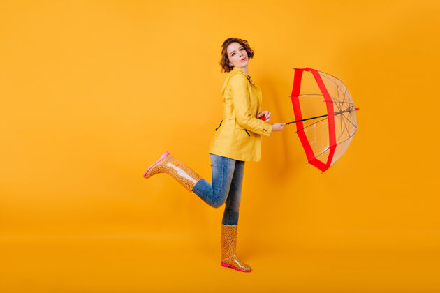 季节穿着橡胶鞋的漂亮女孩和红色阳伞跳舞的全长肖像穿着黄色夹克的卷发女士单腿站着 打着伞室内年轻表情