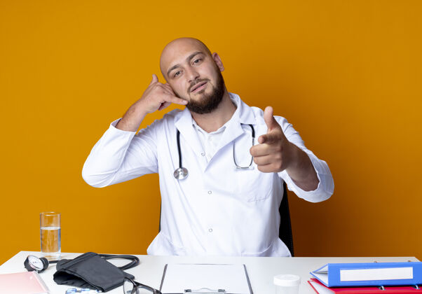 医疗年轻的秃头男医生穿着医用长袍和听诊器坐在办公桌旁 手持医疗工具 显示打电话的手势 指着橙色的摄像头秃头工作橙色