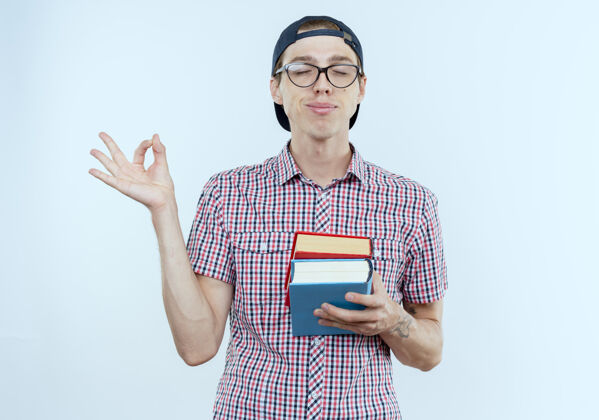 男孩那个戴着背囊 眼镜和帽子 拿着书 在白纸上摆出一副很好的姿势的年轻学生男孩高兴地闭着眼睛背部眼睛学生