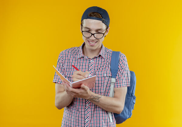 眼镜带着微笑的年轻学生男孩 背着书包 戴着眼镜和帽子 在白色的笔记本上写着什么年轻微笑笔记本