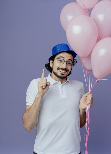 向上笑容可掬的帅哥戴着眼镜 戴着蓝色的帽子 手里拿着气球 指着上面孤立的紫色微笑眼镜男人