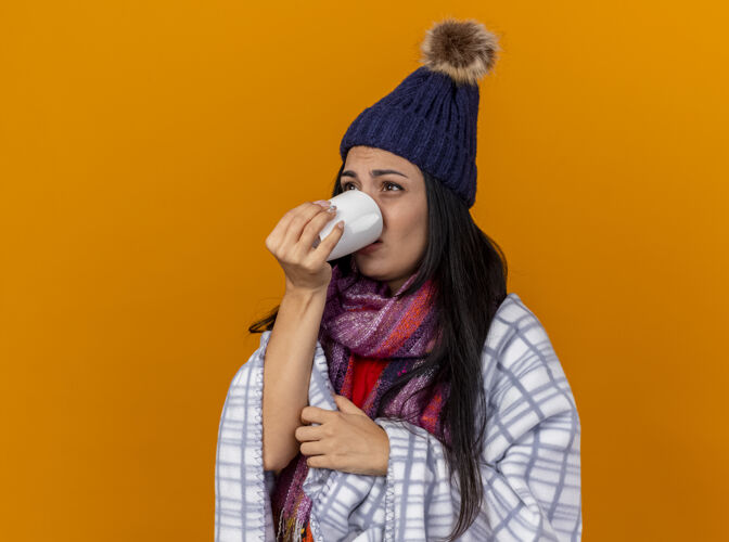女人体弱多病的年轻女子戴着冬天的帽子 围着格子呢围巾站在侧视图里 喝着一杯茶 看起来像是被隔离在橙色的墙上脸穿公民