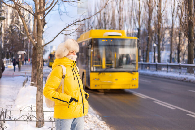 城市一个穿着暖黄色夹克和西伯利亚俄罗斯帽子的快乐女人在雪城街道上等待公共汽车的冬季肖像乐趣公共汽车外套