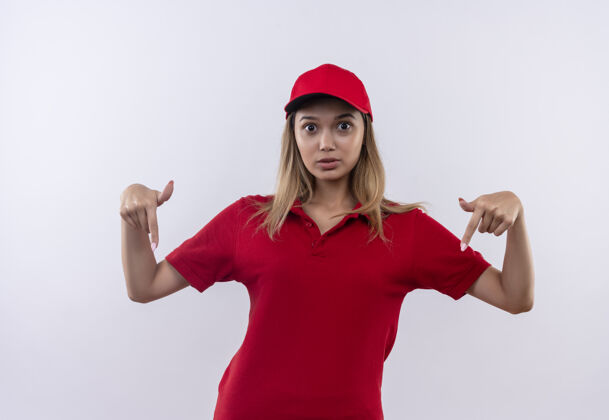 女人惊讶的年轻送货女孩穿着红色制服 戴着帽子点着白色的隔离服年轻手势向下
