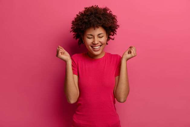 单色快乐女人的形象做拳打手势 庆祝好消息 积极地笑 穿休闲服 积极地笑 对着亮粉色的墙摆姿势情感和成功概念卷发高兴高兴