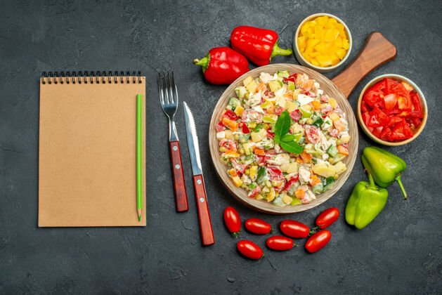 食物深灰色桌子上一碗蔬菜沙拉的顶视图 旁边有餐具 蔬菜和记事本蔬菜桌子美味