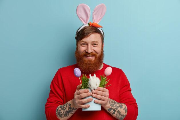 人横拍的快乐姜辣妹表达积极的情绪 戴着兔子耳朵 有纹身 拿着小兔子和两个彩蛋的罐子 象征着复活节节日的概念欧洲事件草