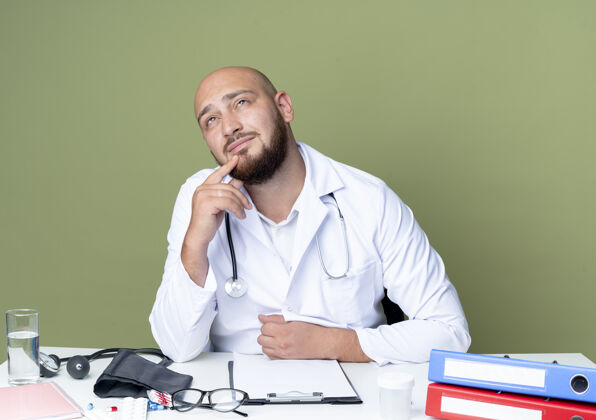 思考想着年轻的秃头男医生穿着医用长袍和听诊器坐在办公桌旁听诊器男性工作