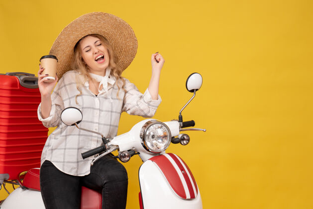 顶戴着帽子坐在摩托车上拿着咖啡享受成功的快乐年轻女子的俯视图人帽子年轻女子