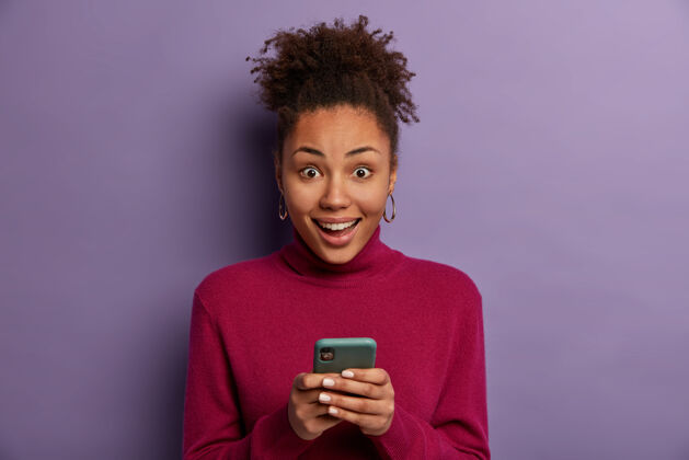 电话喜出望外的卷发女人收到朋友的好消息 拿着手机 发短信 穿休闲套头衫 隔离在紫色的墙上 下载应用程序 浏览互联网青少年女性脸
