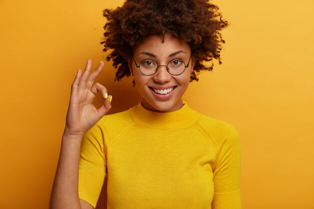 喜悦高兴的卷发女人做了一个可以确认的手势 判断某事 给予认可 说得很好 戴着圆眼镜和t恤 隔离在黄色的墙上 鼓励不要放弃好女性欢呼
