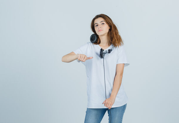 年轻身穿白色t恤 牛仔裤的年轻女性用拇指指着耳机 看上去自信满满 俯瞰前方前面指拇指