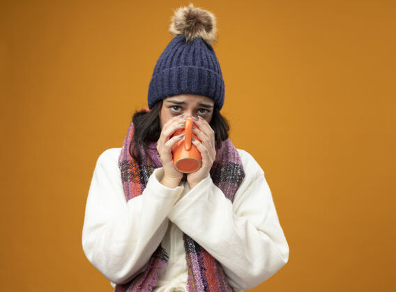 冬天体弱多病的年轻女子戴着长袍 戴着冬帽 戴着围巾 喝着一杯茶 看着前面孤零零的橘黄色墙壁人女人饮酒