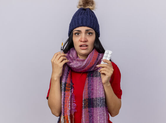 拿着印象深刻的年轻生病的女人戴着冬天的帽子和围巾拿着注射器和一包药片看着前面隔离的白色墙壁围巾冬天姿势