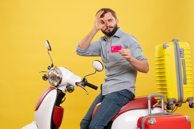 制造旅游概念的正面视图 年轻自信的胡须男子坐在摩托车上 展示黄色的银行卡制作眼镜手势微笑手势成年人