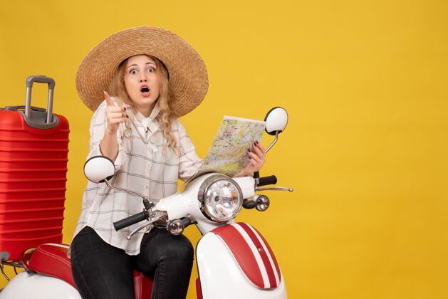 微笑年轻女子戴着帽子 坐在摩托车上 拿着黄色的地图指着前方脸漂亮坐