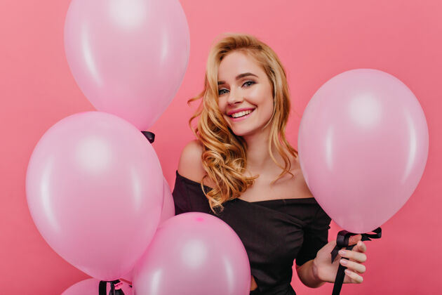 表情无忧无虑的白人女孩带着真诚的微笑在粉红色的气球旁摆姿势室内照片是一位可爱的年轻女子 卷发庆祝生日玩耍明亮服饰
