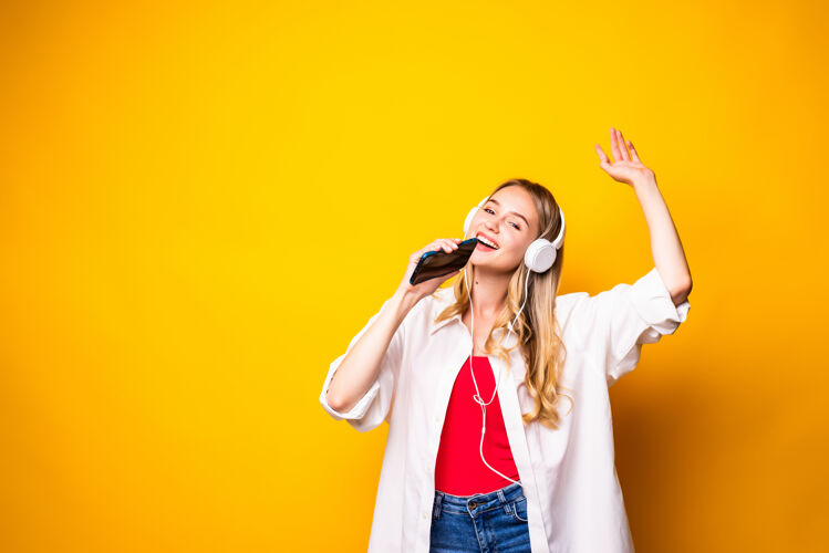 手持微笑的年轻女子戴着耳机听音乐 用智能手机隔着黄色的墙耳机年轻人