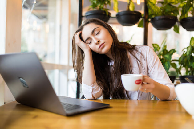 女人一个疲惫的年轻女商人坐在桌边拿着笔记本电脑 一边拿着一杯咖啡 一边在咖啡馆睡觉睡眠休闲休息