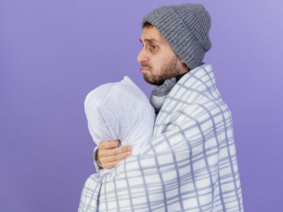 疾病看着身边悲伤的年轻病夫戴着冬天的帽子 围巾裹着格子布 抱住了紫色背景上孤立的枕头拥抱围巾枕头