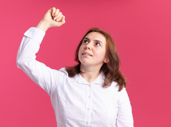 女人身穿白衬衫的年轻女子抬起头 自信地举起拳头站在粉红色的墙上衬衫拳头表情