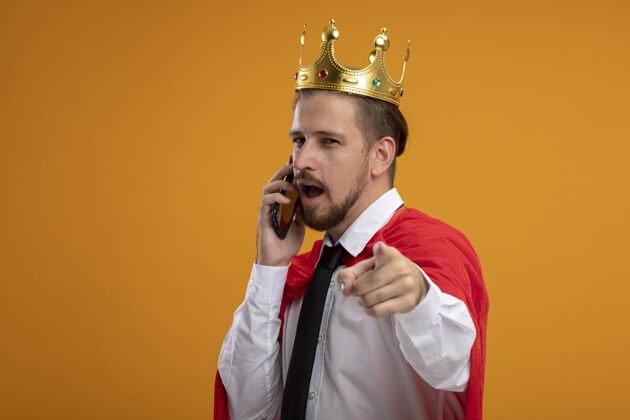 皇冠年轻的超级英雄戴着领带和皇冠 看着摄像机在电话里说话 在橙色的背景下显示出你们孤立的姿态小伙子电话领带