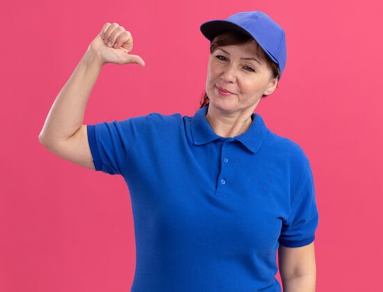 表情身穿蓝色制服 头戴鸭舌帽的中年女送货员看着前面 自信地指着自己站在粉红色的墙上女人年龄中年