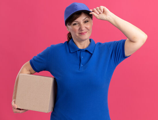 年龄快乐的中年女送货员穿着蓝色制服 戴着帽子 拿着纸板箱看着前面 微笑着自信地站在粉红色的墙上女人微笑抱着